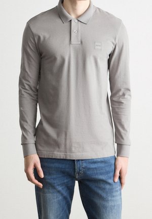 Mann trägt ein graues Langarm-Poloshirt mit zwei Knöpfen und blaue Jeans vor einem schlichten Hintergrund.