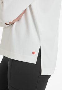 Person wearing white long-sleeve top with a small red embroidered logo near the hem and black fitted pants, hand resting on waist.