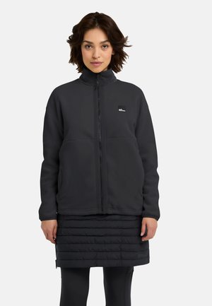 Schwarze Fleecejacke mit hohem Kragen und Frontreißverschluss. Mit einem Logopatch versehen, getragen über einem gesteppte schwarzen Rock und schwarzen Leggings.