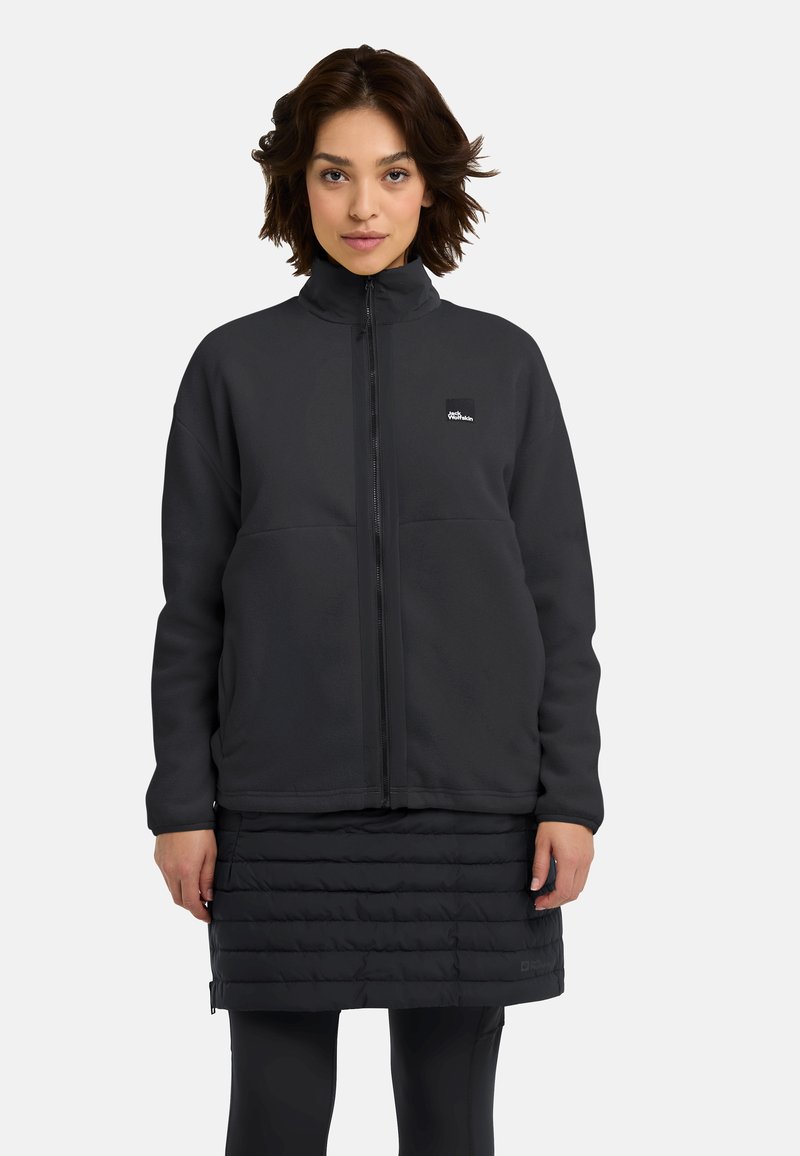 Giacca in pile nera con colletto alto e zip frontale. Presenta una toppa con logo, indossata sopra una gonna trapuntata nera e leggings neri.