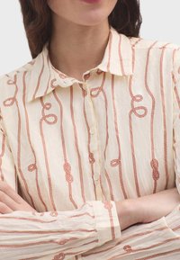 Camisa crema con un patrón de cuerdas rojas verticales, que presenta un cuello clásico y cierre de botones. Tejido texturizado con un aspecto arrugado.