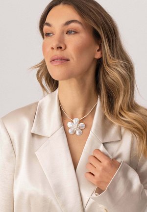 Frau mit welligem blondem Haar, die einen cremefarbenen Satinblazer und eine Halskette mit großem weißem Blumenanhänger trägt und das Revers des Blazers hält.