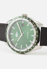 Montre Lacoste avec un cadran analogique vert, un boîtier en acier inoxydable argenté, une fonction date, et un bracelet en cuir noir avec une lunette tachymétrique.