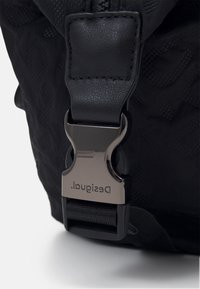 Svart väska med en strukturerad tyg med logotypmönster, framhävd av en metallspänne. Spännet har ingraverad branding och en säker låsning.