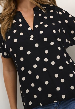 Vrouw met zwarte V-hals blouse met witte stippen en blauwe spijkerbroek, zichtbaar van nek tot heupen tegen een effen achtergrond.