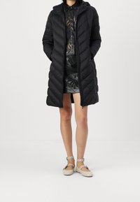 Abrigo puffer negro acolchado con capucha, que cuenta con cremallera frontal y bolsillos laterales. Llevado sobre un vestido negro con estampado y zapatos planos de color claro.