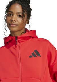 Frau mit lockigem Haar, die eine leuchtend rote Kapuzenjacke mit einem schwarzen Adidas-Logo auf der Brust trägt, vor einem weißen Hintergrund.