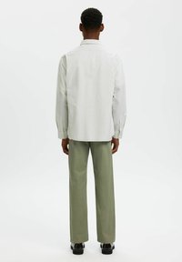 Λευκό πουκάμισο με ρίγες, με γιακά, μακριά μανίκια και κουμπιά στους καρπούς, συνδυασμένο με χαλαρές ελαιόχρωμες παντελόνες και μαύρα παπούτσια.