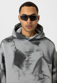Grijze hoodie met een tie-dye patroon, voorzijde met bedrukking en tekstdetails, en een grote capuchon. Model draagt een paar zwarte zonnebril.