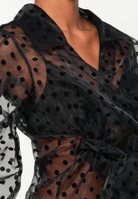 Blouse noire transparente avec des pois noirs, dotée d'un col, de manches longues et d'un détail à nouer à la taille. Texture lisse.