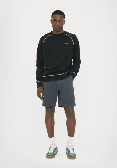 Svart sweatshirt med vit söm och smiley-logotyp, grå shorts och gröna sneakers med vita sulor, bärs med vita strumpor.
