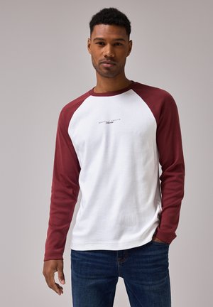 Camicia bianca a maniche lunghe con maniche raglan bordeaux, con testo sul petto. Tessuto morbido in cotone con una vestibilità classica. Indossata con jeans blu.