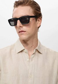 Gafas de sol rectangulares negras con montura de plástico grueso y lentes oscuros, llevadas con una camisa de botones beige claro que presenta un cuello bien definido.