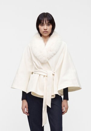 Femme portant un manteau blanc portefeuille avec un grand col duveteux et de larges manches, noué à la taille, associé à un pantalon foncé.
