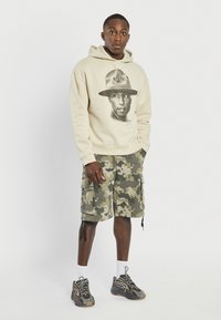 Beige hoodie med et sort grafisk print af et ansigt, parret med grønne camouflagedragt shorts og grå sneakers. Hvide sokker synlige.