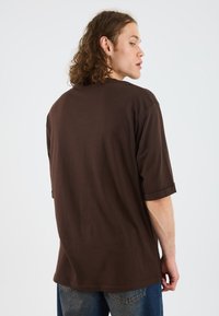 Camiseta de algodón marrón con un corte relajado, mangas cortas enrolladas y cuello redondo, mostrada desde un lado y desde atrás sobre un fondo blanco.