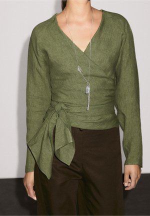 Woman wearing olive green wrap blouse with long tied sash and silver pendant necklace, paired with dark brown pants.