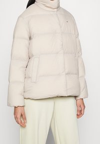 Beige vadderad jacka med hög krage, horisontell quiltad mönstring och sidofickor; tillverkad av ett mjukt, lätt material.