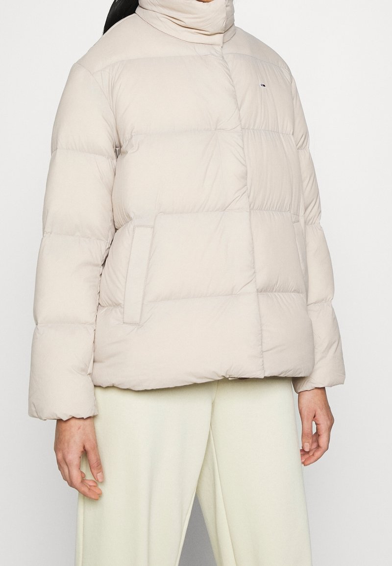 Beige vadderad jacka med hög krage, horisontell quiltad mönstring och sidofickor; tillverkad av ett mjukt, lätt material.