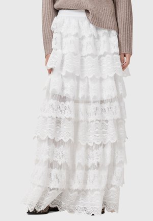 Jupe blanche en dentelle à volants portée avec un pull beige côtelé à manches longues et des chaussures noires visibles à l'ourlet.