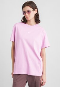 Noor inimene kannab laia heleda lillat Nike T-särki, pruune pükse ja lillat tooni päikeseprille, seistes halli tausta ees.