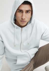 Lys blå hettegenser med glidelås, med snøring i hetten, myk tekstur, og to sidelommer, kombinert med beige bukser.