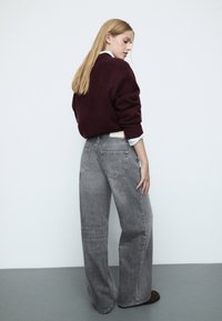 Burgundski oversized pulover uparen s visokim strukom, širokim svijetlo sivim trapericama. Traperice imaju dvije stražnje džepove i laganu teksturu materijala.