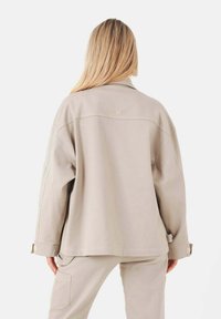 Ljusbeige översized jacka i texturerat tyg med notched krage och uppvikta ärmar, med subtil branding på baksidan.