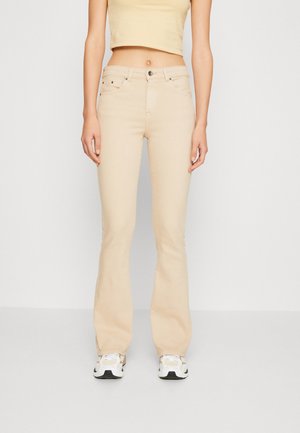 Kobieta w beżowych dzwonach z wysokim stanem, krótkim bezrękawnym topie i białych tenisówkach, stojąca na tle jasnego, jednolitego tła.