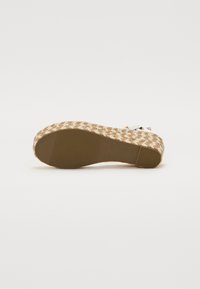 Semelle plate d'une sandale compensée tressée beige avec une semelle en caoutchouc et une petite boucle métallique visible sur la bride.