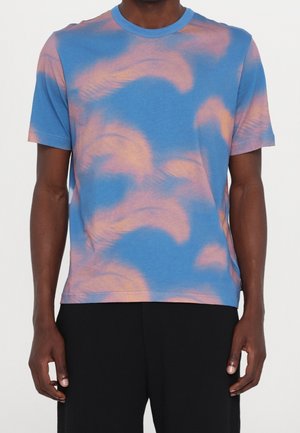 Mężczyzna w niebieskiej koszulce z różowymi, abstrakcyjnymi wzorami przypominającymi pióra oraz w czarnych spodniach, stojący na białym tle.