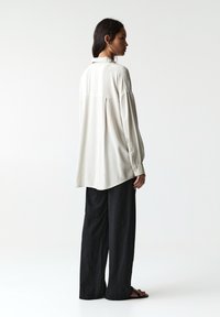 Chemise à manches longues, coupe décontractée, en tissu beige clair avec un pli au dos, assortie à un pantalon noir ample et des sandales plates.