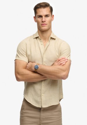 Lichtbeige overhemd met korte mouwen en een button-down kraag; zachte textuur. Gecombineerd met een marineblauw horloge met een tan gekleurde band, poseert het model met gekruiste armen.