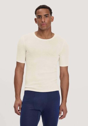 Mann steht vor einem einfarbigen Hintergrund, trägt ein eng anliegendes weißes Kurzarmhemd und dunkelblaue Hose, schaut mit neutralem Gesichtsausdruck nach vorne.