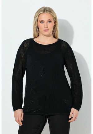 Blondynka w czarnym swetrze z siateczki z długim rękawem i czarnych spodniach, stojąca przy białej ścianie.