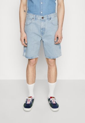 Persona vestida con pantalones cortos de mezclilla azul claro, calcetines blancos tipo crew y zapatillas deportivas azul oscuro con cordones blancos, de pie frente a un fondo liso.