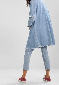 Abrigo de denim azul claro con dobladillo deshilachado combinado con jeans azul claro y zapatillas rojas. El abrigo presenta mangas largas y un ajuste holgado.