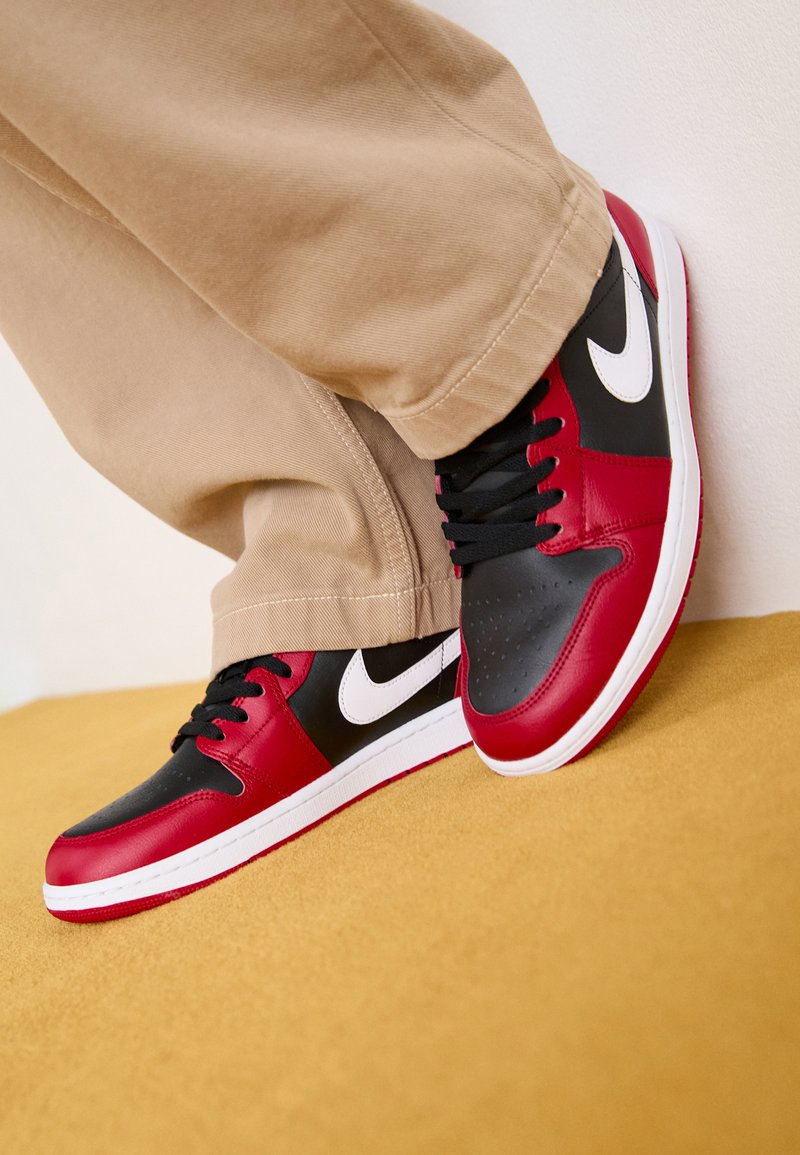 Sneaker in pelle rossa e nera con dettagli bianchi, caratterizzate da un design a bassa tomaia, punta rotonda e lacci neri, posizionate su una superficie gialla.