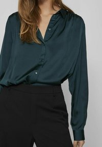 Blouse en soie vert foncé avec une coupe décontractée, manches longues et un col en V à boutons, associée à un pantalon noir ajusté.