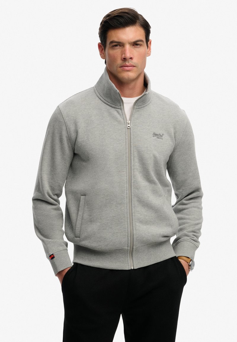 Homme portant une veste zippée gris clair avec des poches et un pantalon noir, debout les mains dans les poches devant un fond uni.