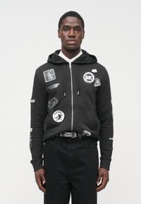 Μαύρο φούτερ με φερμουάρ, εφαρμοστή σχεδίαση, με πολλά μπαλώματα και λογότυπα στο μπροστινό μέρος και στα μανίκια. Υλικό από βαμβάκι, τσέπη καγκουρό, με κουκούλα.