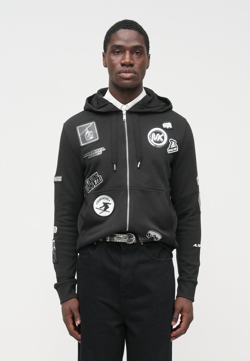 Μαύρο φούτερ με φερμουάρ, εφαρμοστή σχεδίαση, με πολλά μπαλώματα και λογότυπα στο μπροστινό μέρος και στα μανίκια. Υλικό από βαμβάκι, τσέπη καγκουρό, με κουκούλα.