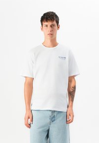 White cotton t-shirt with short sleeves, round neckline, featuring blue embroidered text on the left chest area. Light denim pants.