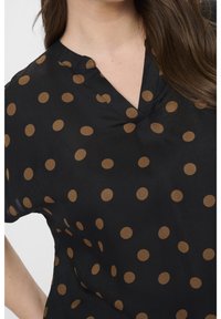 Blouse noire avec un col en V, ornée de pois marron répartis sur tout le tissu. Fabriquée en matière légère, texture douce.