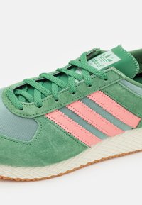 Green athletic shoe with pink stripes, suede and mesh materials, textured light-colored sole, and prominent branding on the tongue.