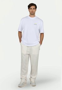 T-shirt branca de manga curta com "PEGADOR" impresso em azul. Combinada com calças texturizadas em creme claro e ténis brancos. Fundo neutro.