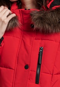 Rode gewatteerde jas met een met bont gevoerd kraag, voorzien van een ritszak en zwarte knopen. Het materiaal lijkt zacht met een doorstikte textuur.