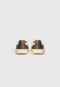 Une paire de baskets basses avec des talons marron, des panneaux supérieurs blanc cassé et des semelles beige clair, montrée de l'arrière sur un fond uni.