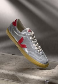Silver athletic shoe with red accents, textured synthetic upper, white laces, and gum rubber sole. Features a prominent 'V' logo on the side.