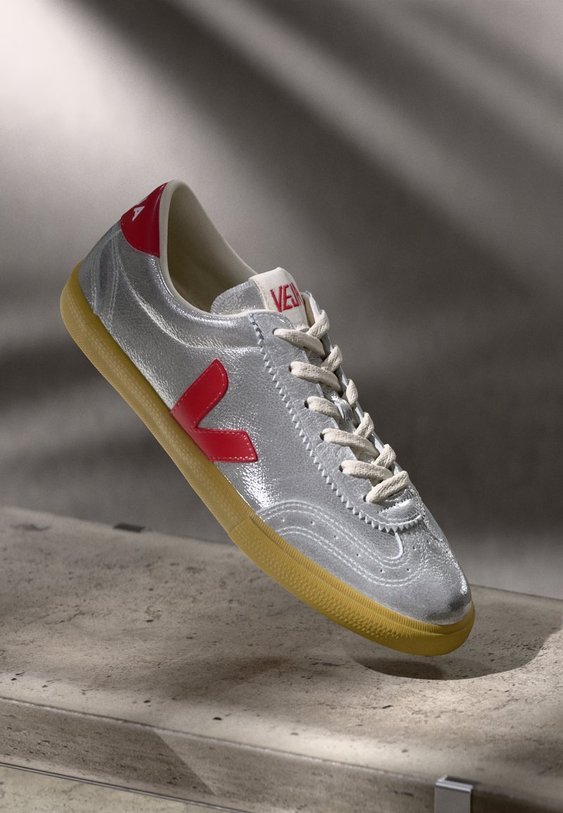 Silver athletic shoe with red accents, textured synthetic upper, white laces, and gum rubber sole. Features a prominent 'V' logo on the side.